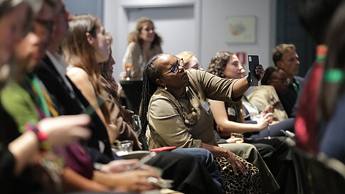 Several people sit in a room, a woman wearing glasses holds up a smartphone to take a photo or video.