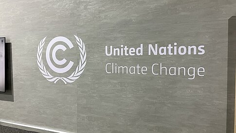 Eine graue Wand mit dem Logo der Klimarahmenkonvention der Vereinten Nationen (UNFCCC). Das Logo besteht aus einem stilisierten „C“ in einem Lorbeerkranz. Rechts daneben steht in weißer Schrift „United Nations Climate Change“.