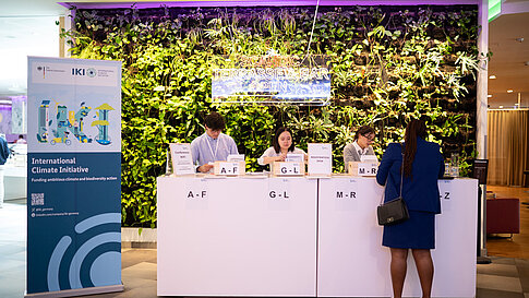Teilnehmer registrieren sich an einem Empfangstisch der Global NDC Conference, der alphabetisch in Abschnitte unterteilt ist. Im Hintergrund befindet sich eine vertikale grüne Pflanzenwand mit einem leuchtenden Schild „Scandic Terrasse Bar Open“. Links steht ein Roll-up der Internationalen Klimaschutzinitiative (IKI).