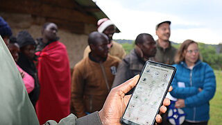 Hand holding smartphone with visible screen in front of a group of people outdoors