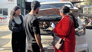 Drei Personen stehen vor einem weißen Auto mit geöffneter Motorhaube, eine Frau in rotem Mantel hält ein Smartphone und zeigt es den anderen beiden.