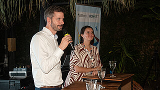 Two people stand at a podium. A man holds a microphone and is speaking, while the person beside him listens attentively. Both look toward the audience. Behind them a banner of the International Climate Protection Initiative is displayed. Full water glasses sit on the tables in front of the speakers.