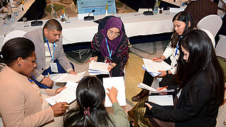 Teilnehmende einer Verhandlungssimulation sitzen mit Papieren und Stiften im Kreis und debattieren.