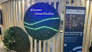 Rundes Schild mit der Aufschrift '#German Climate Pavllion' in orange, daneben ein blauer Infoständer zum IKI-Fotowettbewerb 'Focus. On Climate Action.'