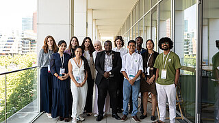 A diverse group of young adults poses for a group photo on a balcony during the Global NDC Conference 2025. They are wearing conference badges and stand in front of a modern building with glass walls.