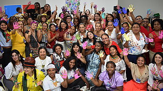 Group of women of various ages and ethnic backgrounds standing in a room showing their hands painted in different colors to the camera
