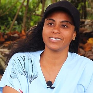 Woman with dark hair wearing a black cap and light blue T-shirt sitting in front of trees and roots speaking into a microphone clipped to her shirt