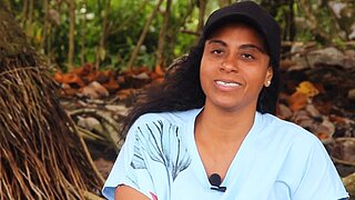 Woman with dark hair wearing a black cap and light blue T-shirt sitting in front of trees and roots speaking into a microphone clipped to her shirt