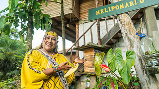 Frau in traditioneller Kleidung steht neben einem Baum mit einem Schild 'MELIPONARIO' und hält ein Bienenstock-Element
