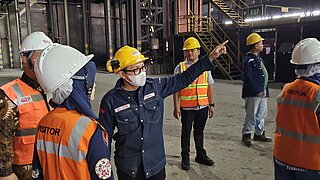 Gruppe von PErsonen mit Schutzhelmen und Warnwesten in einer Industriehalle, eine Person zeigt mit ausgestrecktem Arm nach rechts