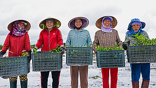 A group of women standing with baskets of plants, smiling while participating in a restoring project in Viet Nam.