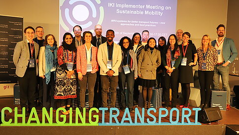 A group of around twenty people poses together on a stage in front of a presentation slide reading “Sustainable Mobility” and the logo “Changing Transport.”