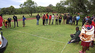 Group of people standing in a circle on grass with one person sitting on the ground holding a rope attached to a car