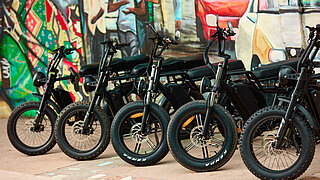 Fünf schwarze Elektro-Lastenräder stehen vor einer bunt bemalten Wand.