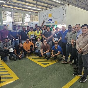A group of people stands in front of a container. They are dressed in work clothes and smiling at the camera. Some are standing, others kneeling. The background shows a banner and a container.
