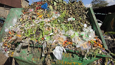 Großer Müllhaufen mit überwiegend organischen Abfällen und Plastikverpackungen in einem grünen Container auf einem Marktgelände