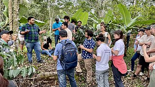 A group of people is standing outdoors in a forested area, listening to a man who appears to be explaining something.