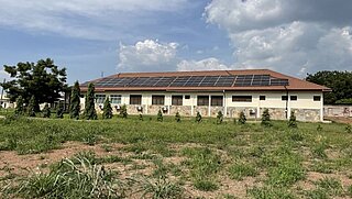 A single-story building with a brown roof covered in solar panels. In front of the building, small trees and grass grow in an open field, with a partially cloudy sky in the background.