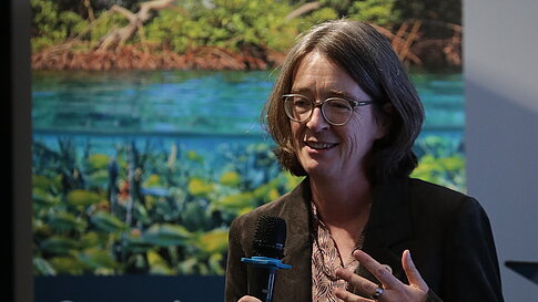 Woman with glasses and dark blazer holding a microphone speaking in front of a banner with nature imagery and text about biological diversity