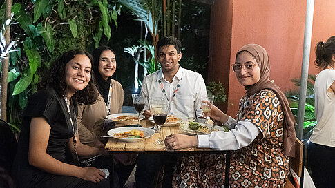 Four participants sit at a wooden table, smiling at the camera, with empty plates and glasses on the table. Tropical plants are visible in the background.