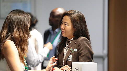 Two women talking at a table with two other people blurred in the background.