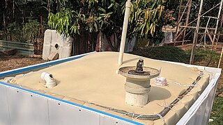 Ein rechteckiger Biodigester mit beige bedeckter Oberfläche und mehreren Rohren steht im Freien vor Bäumen und Metallstrukturen.