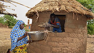 Woman wearing a blue headscarf and patterned skirt holds a metal bowl at a round mud house with a thatched roof where a man in a blue shirt and red scarf passes branches through the window