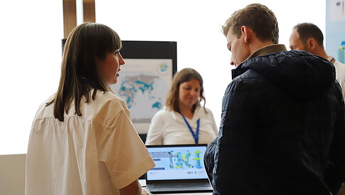 [Translate to English:] Personen stehen an einem Infostand und unterhalten sich.