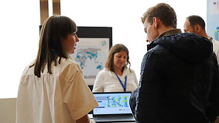 [Translate to English:] Personen stehen an einem Infostand und unterhalten sich.