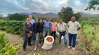In der Bildmitte steht ein Sack mit Kaffeekirschen, während die Gruppe von acht Personen auf einem Kaffeefeld vor einer malerischen Landschaft posiert.