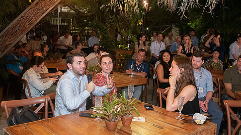Photo of the audience: In the foreground a participant is seated, holding a microphone and gesturing. The gazes of the other seated attendees are directed toward him. Glasses are on the wooden tables. The setting is dark but illuminated, with green plants surrounding the area.