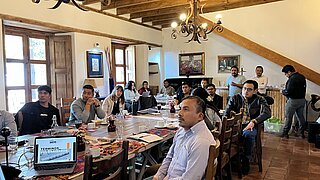 Gruppe von Menschen sitzt an einem langen Holztisch in einem Raum mit sichtbaren Holzbalken und Kerzenleuchtern an der Decke, einige blicken auf einen Laptop mit sichtbarem Text 'TÉRMINOS PULGADOS'