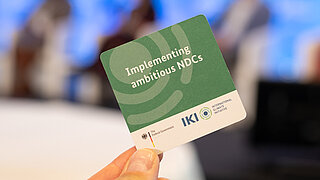 Nahaufnahme einer Hand, die eine grüne Karte mit dem Logo der Internationalen Klimaschutzinitiative (IKI) hält.