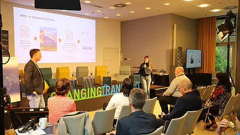 A conference room with several participants watching a presentation. A speaker stands at the front beside a screen displaying slides.
