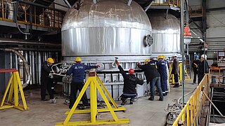 Arbeiter bei der Wartung und Reparatur einer industriellen Anlage in einem großen Produktionsbereich. Sie arbeiten zusammen an einem Gerät, das durch gelbe Sicherheitsbarrieren abgesichert ist.