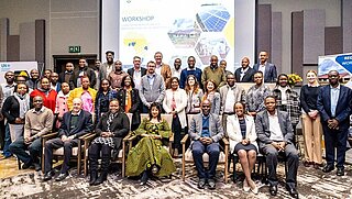 A large group of people poses for a group photo during a regional workshop. Behind them, a presentation screen displays graphics about renewable energy and workshop logos.