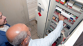 Two men are examining and working on an open electrical panel with various components and wiring.