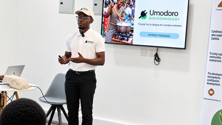 Ein junger Unternehmer in Sambia trägt ein weißes Polohemd und eine weiße Kappe mit dem Logo von Umodoro Biotechnology und präsentiert vor einem Bildschirm mit einer PowerPoint-Folie zu Biogas für den sambischen Markt.
