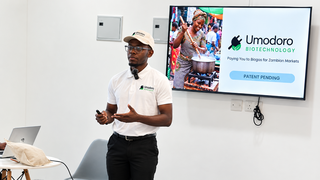 Ein junger Unternehmer in Sambia trägt ein weißes Polohemd und eine weiße Kappe mit dem Logo von Umodoro Biotechnology und präsentiert vor einem Bildschirm mit einer PowerPoint-Folie zu Biogas für den sambischen Markt.