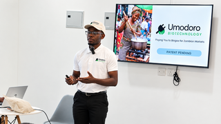 A young entrepreneur in Zambia wearing a white polo shirt and cap with the Umodoro Biotechnology logo presents in front of a screen showing a PowerPoint slide about biogas for the Zambian market.