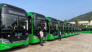 A row of new green and black city buses labeled “DART” parked in a lot, with two people standing in front.