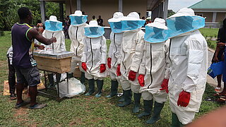 Sieben menschen in Bienenschützanzügen stehen auf einer Wiese vor einem Bienenstock, während ihnen der Umgang mit den Materialien erklärt wird.