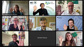 A screenshot of a video conference with several participants. The individuals in each window are waving at the camera. Different people are seen with various backgrounds, including bookshelves, artwork, and plants. Some names are displayed in the middle, with microphone and camera icons visible.