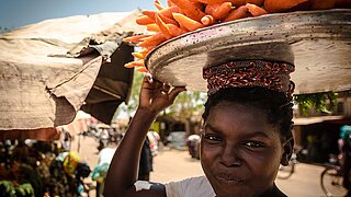 Young woman carrying a large metal tray of carrots on her head holding it with one hand