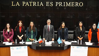 A group of seven people stands behind a table with water bottles and documents. The banner in the background reads "La Patria es Primero."