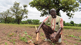 A man kneeling on a field holding a hoe with trees and cloudy sky in the background
