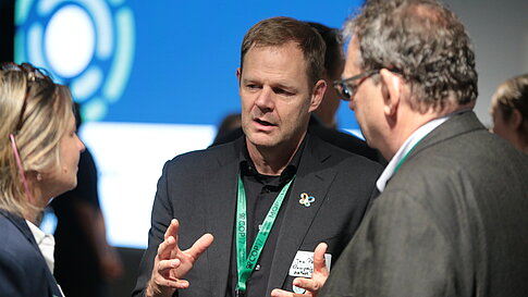 Three people in conversation, one person gesturing with hands, blurred screen with blue design in the background