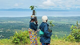 Two women in traditional African dress are carrying two seedlings