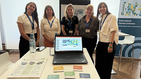 A group of five people stands at a booth for the International Climate Initiative. On the table, informational materials and a laptop displaying a presentation are visible.