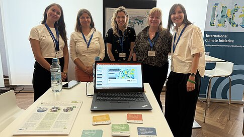 Eine Gruppe von fünf Personen steht an einem Stand der Internationalen Klimaschutzinitiative. Auf dem Tisch sind Informationsmaterialien und ein Laptop sichtbar, der eine Präsentation zeigt.